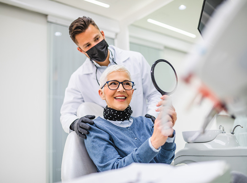 A dentist is assisting an elderly woman with a dental procedure, both in protective face masks.