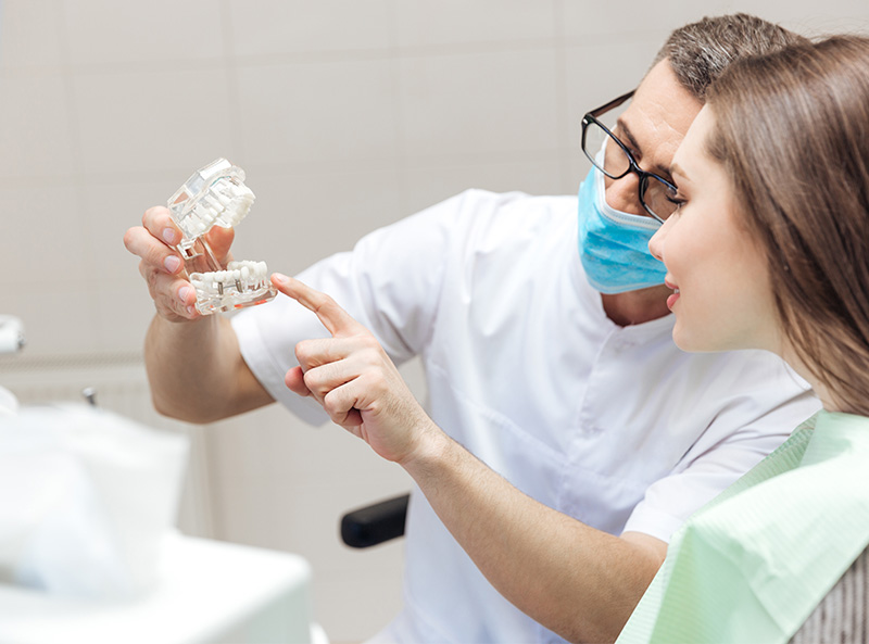 In the image, a dentist in a white coat is examining a patient s teeth with a magnifying glass, while a woman watches attentively.