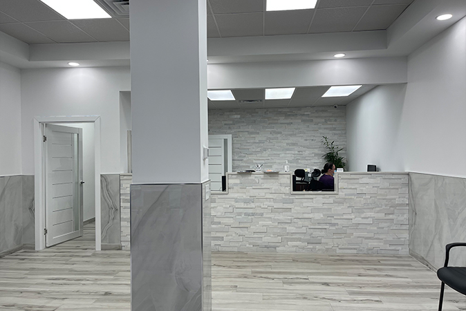The image shows an interior view of a modern office space featuring a reception area with marble countertops, a white wall with a brick-like texture, and a person seated at the desk.