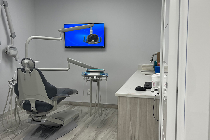 The image depicts a modern dental office with a chair, dental equipment, and a desk with a computer monitor.
