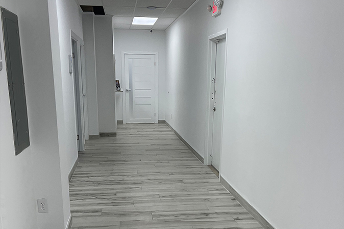 The image shows an interior hallway with a white wall on the left, a door at the end of the corridor, and flooring that appears to be a light-colored wood or laminate.